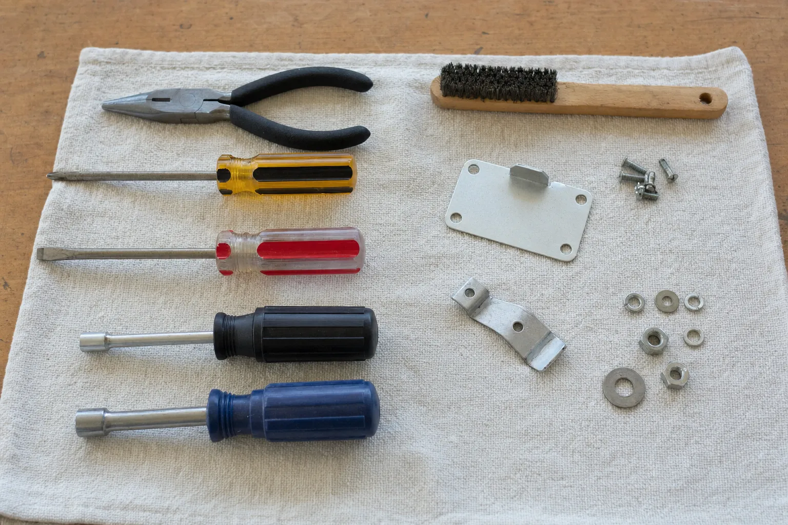 Screwdrivers, nut drivers, pliers, a wire brush and two flat brackets laid out on a canvas cloth
