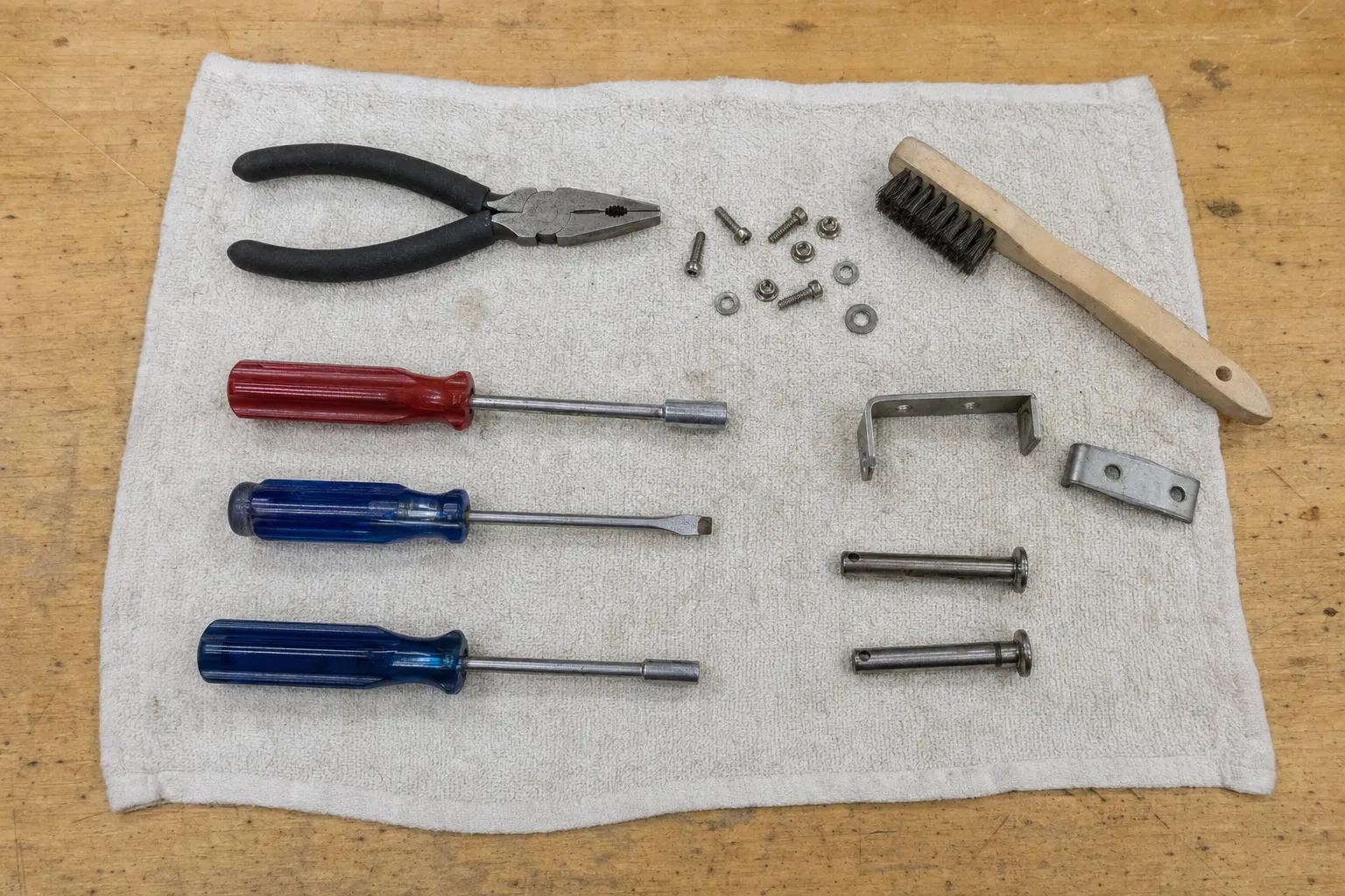 Towel on a wooden bench holding pliers, two nut drivers, a slotted screwdriver and a wire brush beside a U-bracket, a small plate and two steel pins